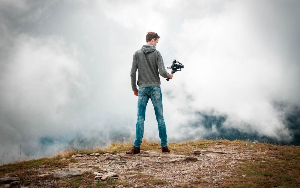 A boy in jeans standing on a rock with a professional camera in his hand filming clouds in front of him.